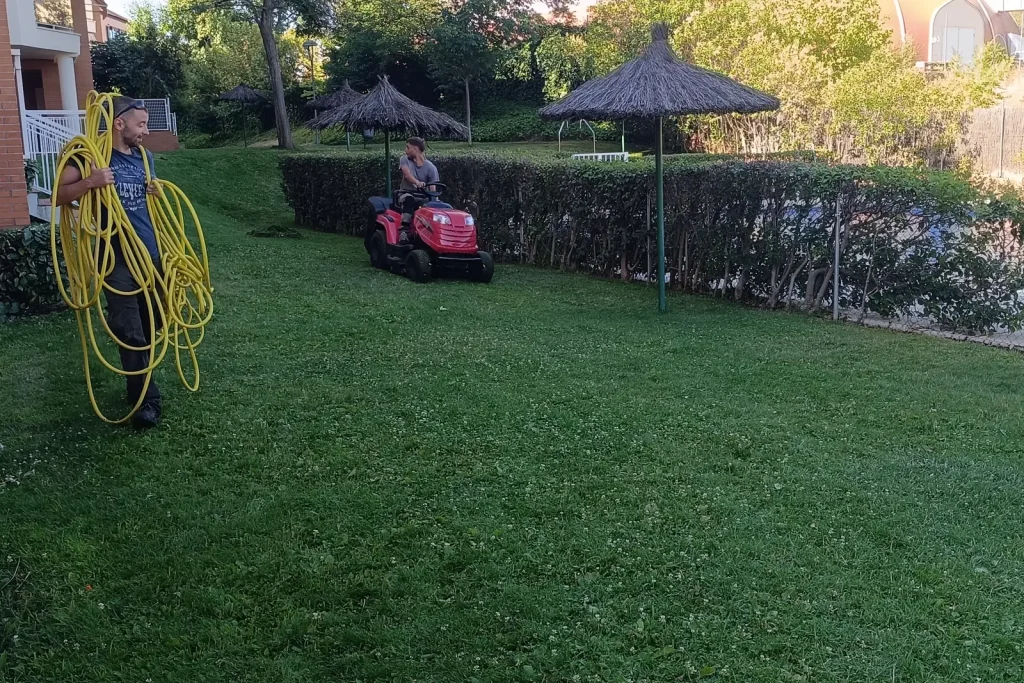 Dos jardineros segando el césped con tractor cortacésped y recogiendo una manguera