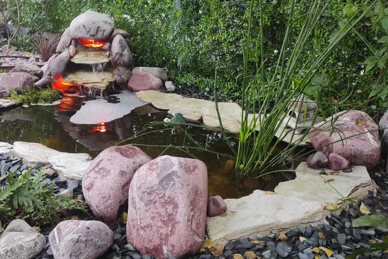 Estanque de jardín con cascada de piedra iluminada, rocas rojizas y plantas acuáticas